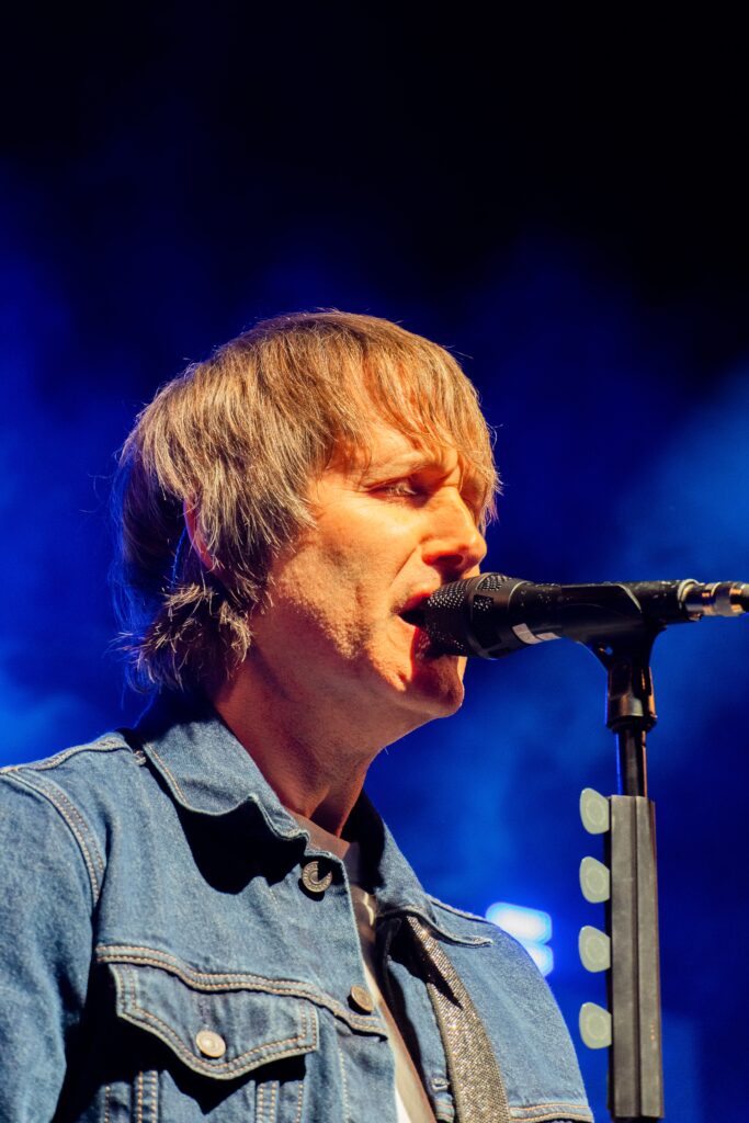 Close-up of a male singer with bangs and a denim jacket singing intensely into a stage microphone.